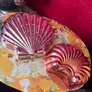 Puede incluir: Dos moldes con forma de concha de color cobre. Uno es una gran concha de vieira y el otro es una concha de nautilus más pequeña. Se encuentran sobre una superficie estampada en amarillo y naranja, con un fondo rojo.