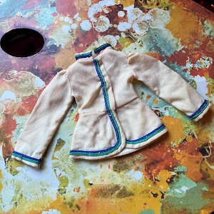 May include: A cream-coloured vintage doll jacket with blue and green trim. The jacket features long sleeves and a high collar. The trim is along the edges and down the front. The jacket is made of a soft, possibly felt-like material.