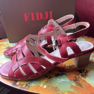 May include: A pair of red leather sandals with a cage-like design. The sandals have a small heel and adjustable ankle straps with silver buckles. The brand name "FIDJI" is visible on the box in the background.