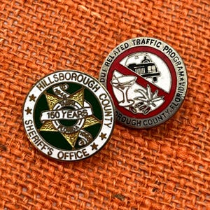 Puede incluir: Dos pines metálicos circulares. Uno presenta una estrella verde y dorada con el texto Hillsborough County Sheriff's Office 150 Years. El segundo tiene un borde rojo con el texto DUI Related Traffic Program Hillsborough County Florida.