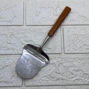 May include: A metal cheese slicer featuring a flat, wide blade with a horizontal cutting slot and a smooth, cylindrical amber-colored handle. The tool has a polished silver-tone finish on the metal portion and a solid, warm-toned handle.