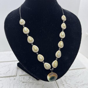 May include: A necklace featuring cream-colored, teardrop-shaped beads with a silver chain. The necklace has a large, round pendant with brown and green hues. The necklace is displayed on a black mannequin.