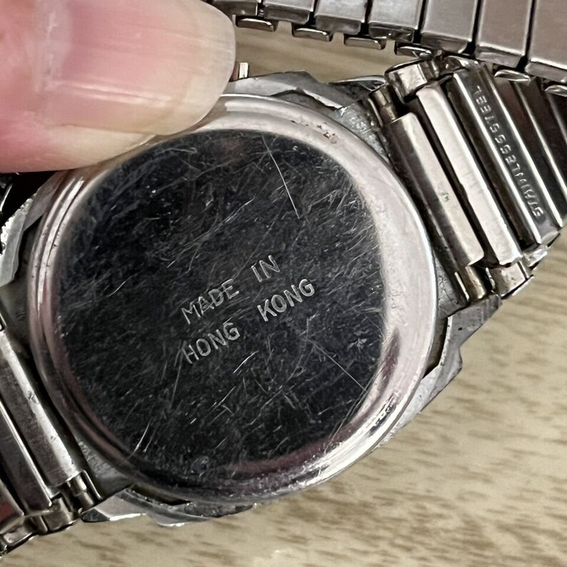 Op de afbeelding: Close-up van een zilverkleurige horlogerug met de tekst "MADE IN HONG KONG" gegraveerd. De horlogeband is van roestvrij staal. Het horloge lijkt vintage te zijn.