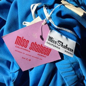 Könnte beinhalten: Nahaufnahme eines leuchtend blauen Kleidungsstücks mit angebrachten Etiketten. Auf dem rosa Etikett steht "miss shaheen CALIFORNIA HAWAII" und "Size 7/8". Ein kleineres Etikett lautet "Miss Shaheen CALIFORNIA HAWAII". Der Stoff ist leuchtend blau.