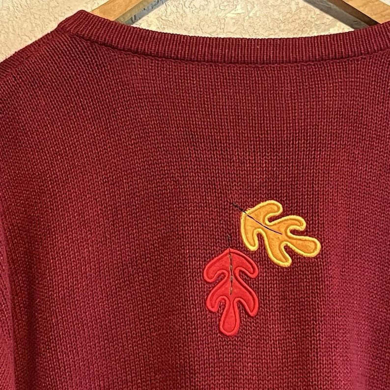 May include: A burgundy knit sweater with two embroidered autumn leaves. One leaf is red with a black outline, and the other is yellow with a black outline. The sweater has a crew neck.