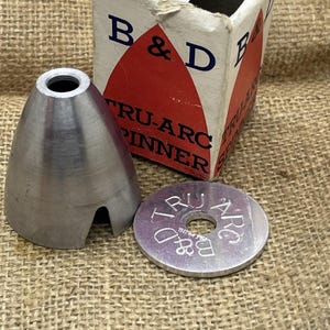 Op de afbeelding: Een vintage Tru-Arc spinner set, met een zilverkleurig kegelvormig stuk en een plat, cirkelvormig stuk met de tekst "Tru-Arc" en "B & D" gegraveerd. De originele doos is zichtbaar op de achtergrond.