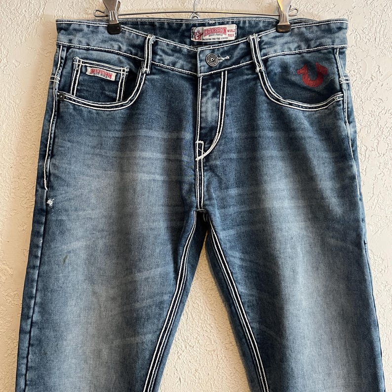 Pu&ograve; includere: Jeans blu denim con cuciture bianche e lavaggio sbiadito. Presentano tasche anteriori e posteriori, chiusura con bottone e logo rosso sulla tasca anteriore. I jeans sono appesi a una gruccia di metallo.