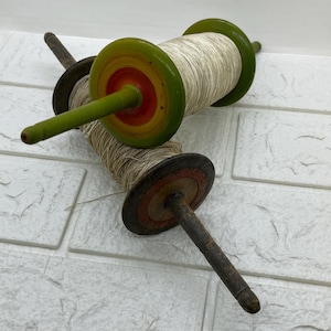 May include: Two vintage wooden kite string winders with green, yellow, and red painted details. The winders are made of wood and have a metal spindle. The string is white and is wound around the winders.