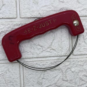 Peut inclure: Un outil rouge "E-Z-JUST" avec une poignée et une boucle en fil de fer. L'outil porte des lettres dorées et un bouton argenté. La poignée est incurvée et porte le petit numéro "61" sur le côté. L'outil est conçu pour un usage spécifique.