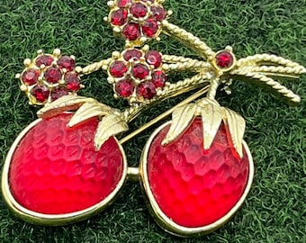 May include: A gold-toned brooch featuring two red strawberry-shaped elements with textured surfaces and gold-colored leaves. The brooch also includes clusters of red gemstones and a gold-colored stem.