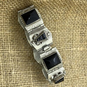 Puede incluir: Pulsera plateada con tres piedras rectangulares de ónix negro. La pulsera tiene un diseño ornamentado con detalles grabados y un cierre de broche. La pulsera se muestra sobre una superficie texturizada de color beige.