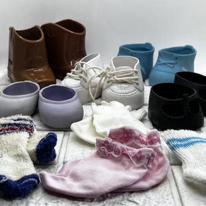 Op de afbeelding: Een verzameling miniatuur poppenschoenen en accessoires, waaronder bruine cowboylaarzen, witte sneakers, blauwe en zwarte schoenen en diverse sokken. De sokken zijn wit, roze en blauw met decoratieve randen. De items zijn gerangschikt op een witte ondergrond.