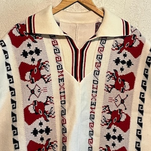 May include: A cream-colored knit pullover featuring a collar with red, white, and black striped trim. The garment displays vertical panels with red deer head motifs, black geometric patterns, and the text MEXICO printed in black.