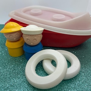 Könnte beinhalten: Ein Vintage-Spielzeug-Bootsset mit einem roten Boot mit rosa Innenraum und weißer Verkleidung, zwei weißen Rettungsringen und zwei Spielfiguren. Eine Figur trägt einen gelben Hut, die andere eine Matrosenmütze. Die Figuren stehen auf blauen und gelben Sockeln.