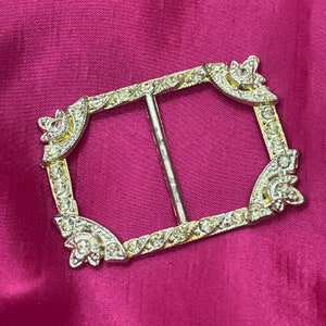 May include: A rectangular silver-toned buckle with clear rhinestones. The buckle has a central bar and decorative floral accents at each corner. The background is a solid magenta color.