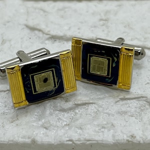 May include: A pair of silver and gold cufflinks. Each cufflink has a dark blue square center with a gold-colored microchip design. The edges are gold with a fluted column design. The cufflinks are on a white and gray surface.