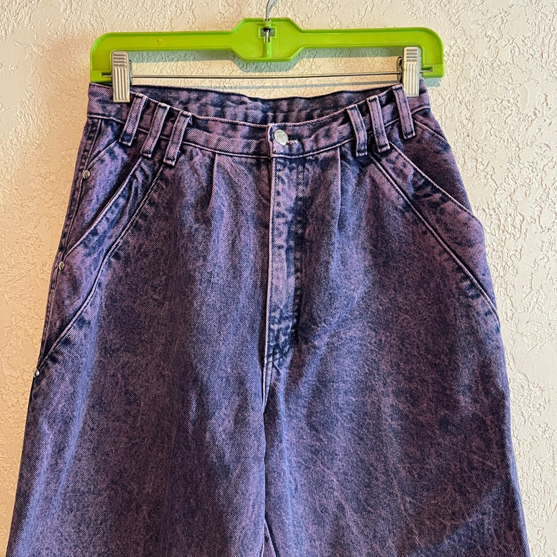 May include: Close-up of acid wash purple denim jeans. The high-waisted jeans have a button and zipper closure, belt loops, and front pockets. The denim has a faded, textured appearance. The jeans are hanging on a green hanger.