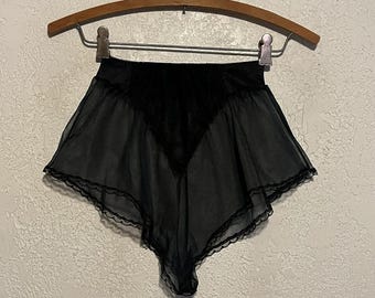 May include: High-waisted black shorts made of a sheer, lightweight fabric. The garment features a solid black opaque panel at the center front, a scalloped lace trim along the bottom hem, and an elasticized waistband, all displayed on a hanger.