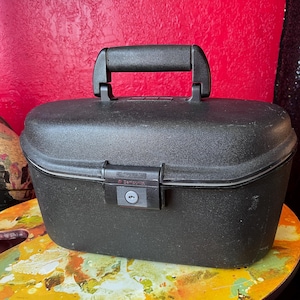 May include: A black Samsonite travel case with a handle and a latch closure. The case is oval-shaped and sits on a colorful surface. The handle is located on the top of the case.