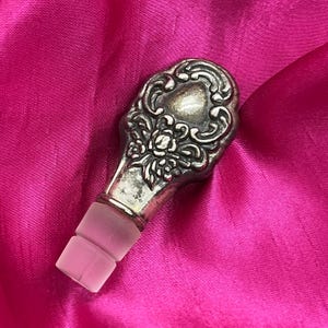 Puede incluir: Tapón de botella plateado con un diseño floral ornamentado. El tapón tiene una base de vidrio esmerilado rosa. La parte superior de metal presenta un detallado patrón de flores y volutas. El fondo es una tela rosa brillante.