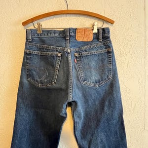 Op de afbeelding: Donkerblauwe denim jeans met een klassiek ontwerp met vijf zakken, hangend aan een houten hanger. De jeans hebben een rood label op de achterzak en een leren patch. De jeans zijn een vintage stijl.