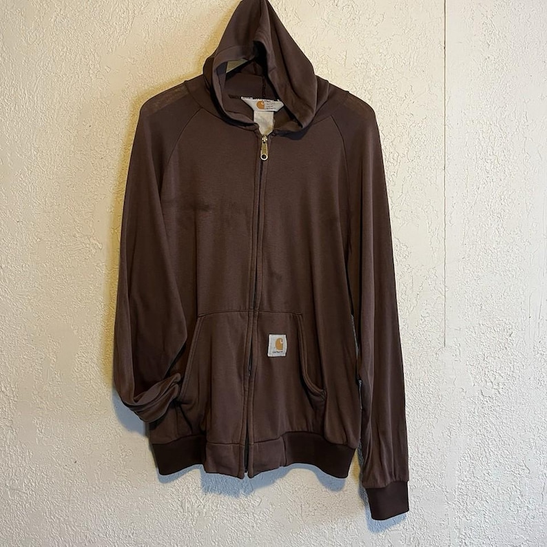 May include: A brown zip-up hoodie with a hood and front pockets. The hoodie has a small Carhartt logo patch on the front pocket and a full-length zipper. The cuffs and bottom hem are a darker shade of brown.