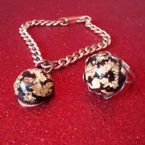 May include: A gold-toned bracelet with a matching ring. The bracelet has a chain link design with a round charm. The ring and charm are black with gold leaf accents. The jewelry set is displayed on a red glitter surface.