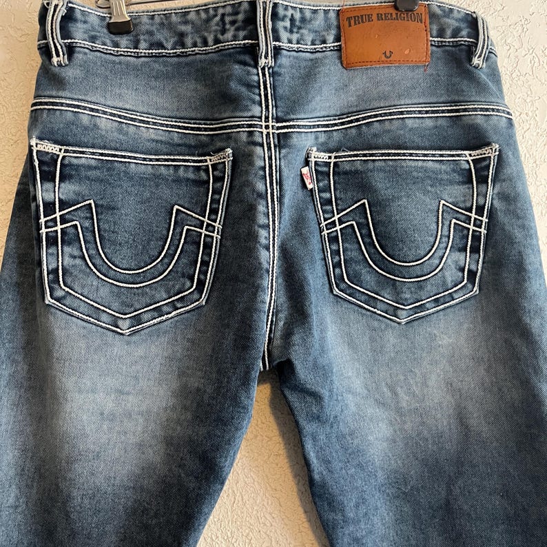 Pu&ograve; includere: Jeans blu in denim con cuciture bianche e design decorativi sulle tasche posteriori. I jeans presentano una toppa in pelle marrone con il logo "True Religion". Le tasche posteriori hanno un esclusivo design a forma di ferro di cavallo.