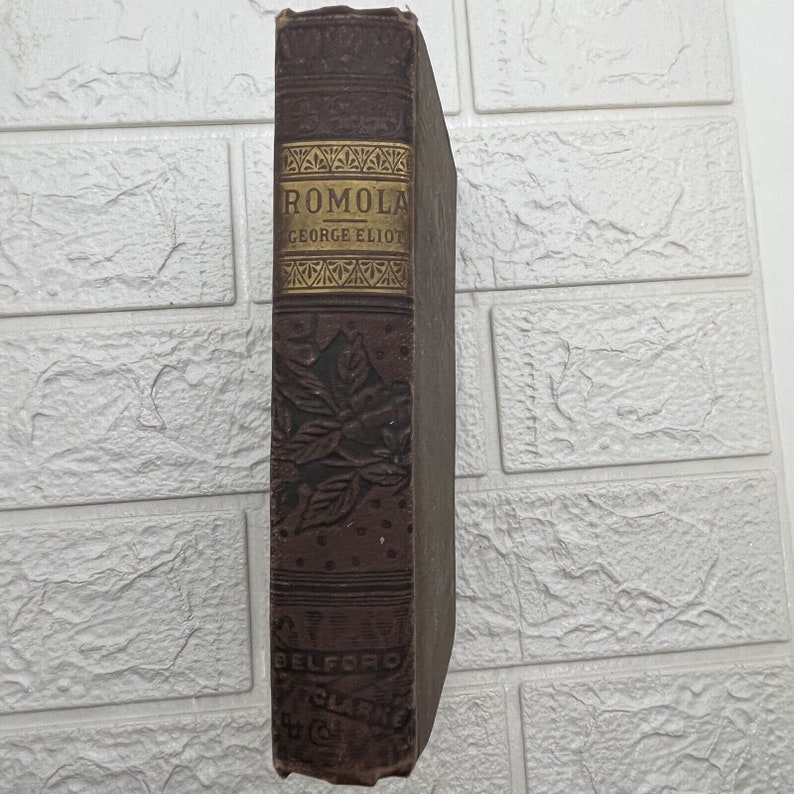 May include: A brown hardcover book with a gold-stamped spine featuring the title Romola by George Eliot. The spine displays embossed floral patterns and the publisher name Belford, Clarke & Co. at the bottom, presented in a vertical orientation.