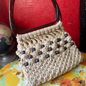 May include: Cream-colored macrame handbag with dark brown wooden beads and a brown plastic handle. The bag has a woven design and a rectangular shape. The handle is curved and attached to the top of the bag.