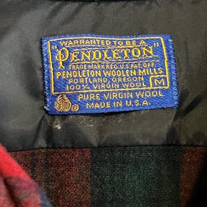 May include: A blue rectangular clothing label with yellow text reading WARRANTED TO BE A PENDLETON, PENDLETON WOOLEN MILLS, PORTLAND, OREGON, 100% VIRGIN WOOL, M, PURE VIRGIN WOOL, MADE IN U.S.A. stitched onto a black lining above a red and green plaid fabric.