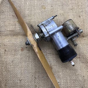 Puede incluir: Un antiguo motor de avión modelo con una hélice de madera. El motor es plateado con un cilindro negro acanalado y una bujía. La hélice es de color marrón claro y está unida al motor.