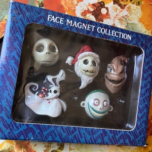 Può includere: Una collezione di cinque calamite a forma di volti dei personaggi del film "Nightmare Before Christmas" in una scatola blu. Le calamite includono Jack Skellington, Zero e altri personaggi. La scatola riporta la scritta "FACE MAGNET COLLECTION".