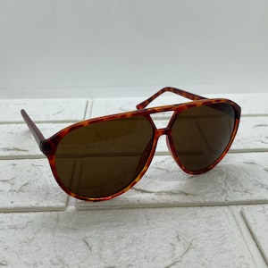 May include: Brown aviator sunglasses with a tortoiseshell pattern frame. The sunglasses have a double bridge and brown tinted lenses. The frame is a mix of orange, brown, and yellow.