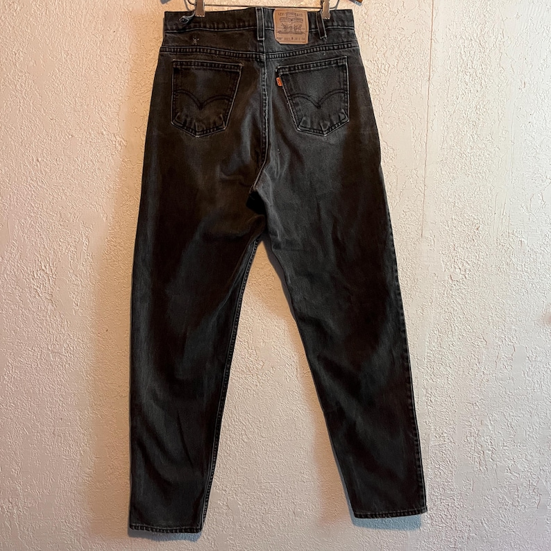 K&ouml;nnte beinhalten: R&uuml;ckansicht einer verblichenen schwarzen Jeans. Die Jeans hat ein klassisches F&uuml;nf-Taschen-Design, mit dem Levi's-Markenaufn&auml;her auf dem Bund. Das orangefarbene Levi's-Etikett befindet sich auf der Ges&auml;&szlig;tasche. Die Jeans h&auml;ngt an einem Kleiderb&uuml;gel.