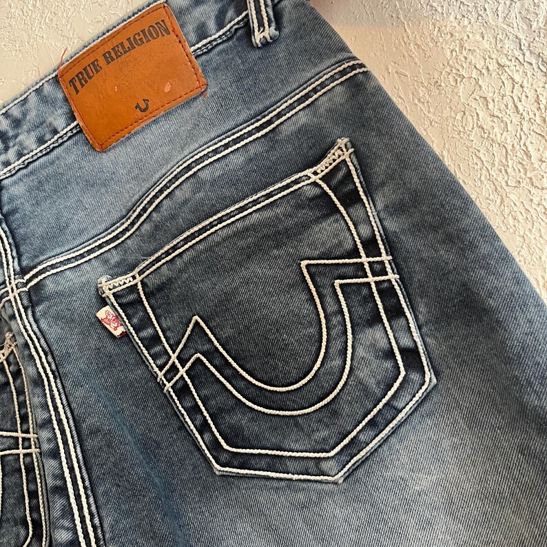 Pu&ograve; includere: Primo piano di jeans blu con cuciture bianche. La tasca posteriore presenta un caratteristico design cucito in bianco. Una toppa in pelle marrone con la scritta "TRUE RELIGION" &egrave; visibile vicino alla cintura. Una piccola etichetta rossa &egrave; attaccata alla tasca.