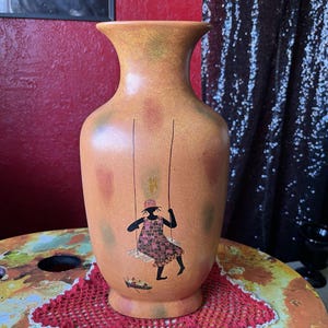 Puede incluir: Un jarrón de cerámica marrón con una superficie texturizada y un diseño pintado de una silueta negra de una persona en un columpio. La persona lleva un vestido estampado en rojo y blanco.