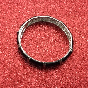 Op de afbeelding: Een zilverkleurige scharnierende armband met zwarte emaille accenten. De armband heeft een geweven ontwerp aan de binnenkant en rechthoekige zwarte emaille secties. De armband is geplaatst tegen een rode glitter achtergrond.
