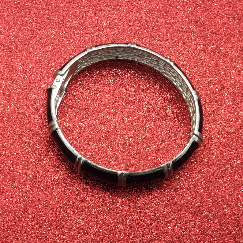 Op de afbeelding: Een zilverkleurige scharnierende armband met zwarte emaille accenten. De armband heeft een geweven ontwerp aan de binnenkant en rechthoekige zwarte emaille secties. De armband is geplaatst tegen een rode glitter achtergrond.