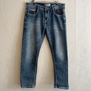 Pu&ograve; includere: Jeans di denim blu con cuciture bianche e un lavaggio sbiadito. I jeans hanno un classico design a cinque tasche, una chiusura con bottone e cerniera e un dettaglio con logo rosso sulla tasca anteriore. I jeans sono appesi a una gruccia di metallo.