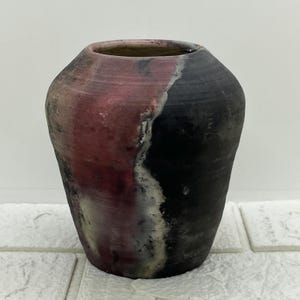 Può includere: Un vaso in ceramica fatto a mano con una combinazione di colori unica. Il vaso presenta una miscela di tonalità rosse, nere e bianche. Il design è diviso diagonalmente, con una sezione rossa da un lato e una sezione nera dall'altro. Il vaso ha un'ampia apertura e una superficie strutturata.