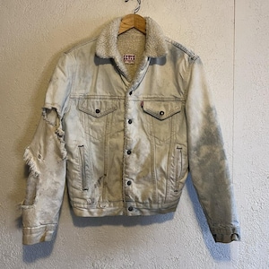 May include: A distressed, light-wash denim jacket with a cream-colored shearling collar and lining. The jacket features button closures, chest pockets, and side pockets. The left sleeve has a large tear. A small red Levi's tag is visible.