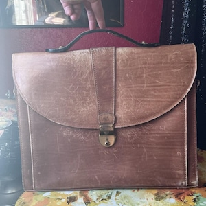 May include: A vintage brown leather briefcase with a flap closure and a handle. The briefcase has a brass-colored lock and shows signs of wear, adding to its classic appeal. The bag is designed for carrying documents and other essentials.