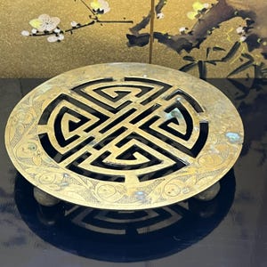 May include: A round, antique brass trivet with a central geometric design and fish motifs around the edge. The trivet has three small feet and a dark background. The brass has a warm, aged patina.