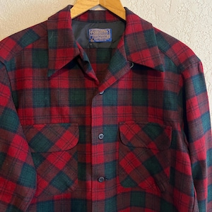 May include: A long-sleeve button-up shirt featuring a red and dark green plaid pattern. The shirt has a pointed collar, two front chest pockets with flaps, and a blue Pendleton brand label sewn into the inner neckline. The fabric appears to be wool.