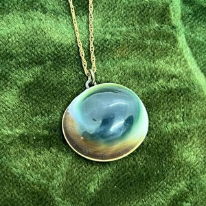 Pode incluir: Um colar dourado com um pendente grande e iridescente. O pendente tem uma forma arredondada com uma borda dourada e apresenta tons de azul, verde e castanho. A corrente tem um design delicado e torcido.