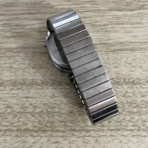 Op de afbeelding: Een zilverkleurig polshorloge met een rekbare metalen band. De wijzerplaat van het horloge is zichtbaar en de band is gedeeltelijk uitgeschoven. Het horloge ligt op een lichtgekleurd houten oppervlak.