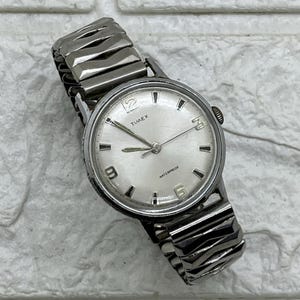 Może przedstawiać: Zegarek na rękę Timex w stylu vintage ze srebrną tarczą i rozciągliwą bransoletą ze stali nierdzewnej. Tarcza zegarka wyświetla nazwę marki, słowo "Waterproof" oraz cyfry 3, 6, 9 i 12. Zegarek ma srebrne wskazówki i znaczniki.