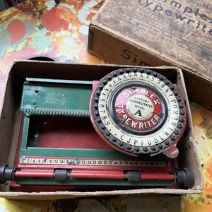May include: A vintage Simplex Typewriter, Model A, in its original cardboard box. The typewriter is red and green with a circular dial for selecting letters and numbers. The box has text that reads "Simplex Typewriter."
