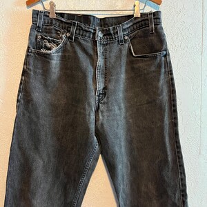 K&ouml;nnte beinhalten: Eine verblasste schwarze Jeans mit klassischem F&uuml;nf-Taschen-Design. Die Jeans hat einen Knopf- und Rei&szlig;verschluss, G&uuml;rtelschlaufen und ein abgenutztes Taschen Detail. Die Jeans h&auml;ngt an einem Holzb&uuml;gel.